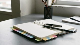 An open ring bound notebook on a gray desk with alphabet tabs on its edge, a pen across the page and arrow shafts standing in a holder behind