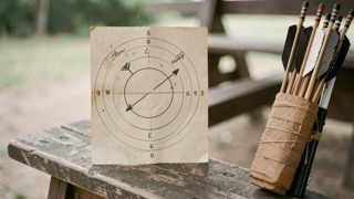 A clipboard score sheet on a wooden fence rail at a sheepdog trial, phase names printed down the left column and pencil marks in the right, late afternoon light across an open field with sheep in the far background.