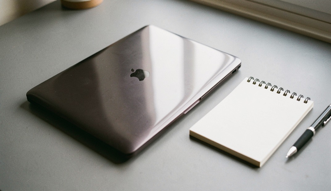 A closed laptop on a grey desk catching a soft window reflection, a blank notepad and a pen squared beside it on the surface