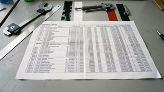 A one design class rule open on a workbench beside a caliper and a roll of arrow wrap film, late afternoon light from a side window, shot from slightly above at a three quarter angle.