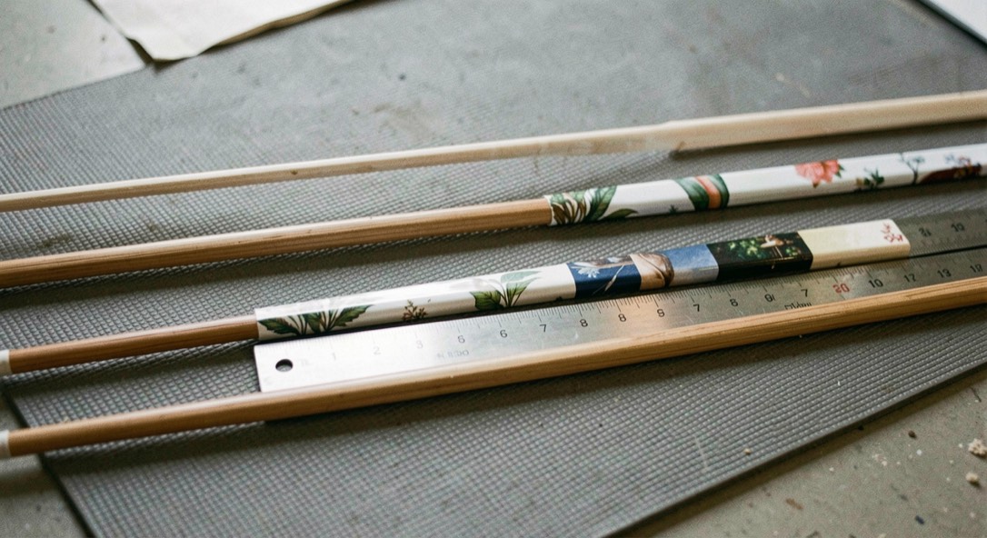 Three arrow shafts laid parallel on a grey mat, each carrying a band of printed vinyl of a different length, a steel rule across them